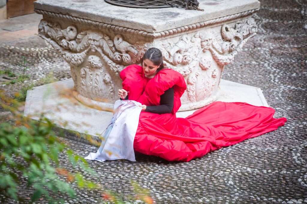 Nel gesto paziente del cucire, Giovanna Zampagni trasforma la fotografia in un territorio attraversabile, dove memoria e presenza si intrecciano. Accanto al pozzo del chiostro, il corpo avvolto nel rosso diventa parte dell’opera, dando forma a una relazione fragile e viva tra immagine, tempo e trasformazione.