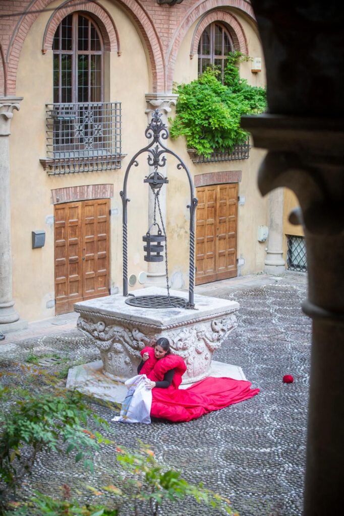 Avvolta nel rosso intenso del mantello, Giovanna Zampagni abita il chiostro come uno spazio sospeso tra memoria e presenza. Il corpo raccolto accanto al pozzo trasforma il gesto performativo in un’immagine silenziosa e fragile, dove il tempo rallenta e il quotidiano lascia emergere i suoi fantasmi.